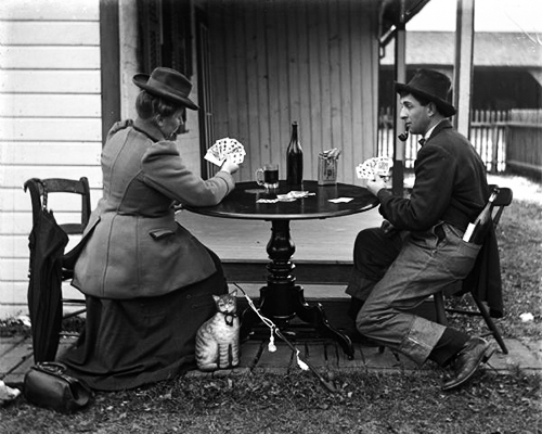 A dapper woman and man play cards at a table outside with a cat looking at the picture taker